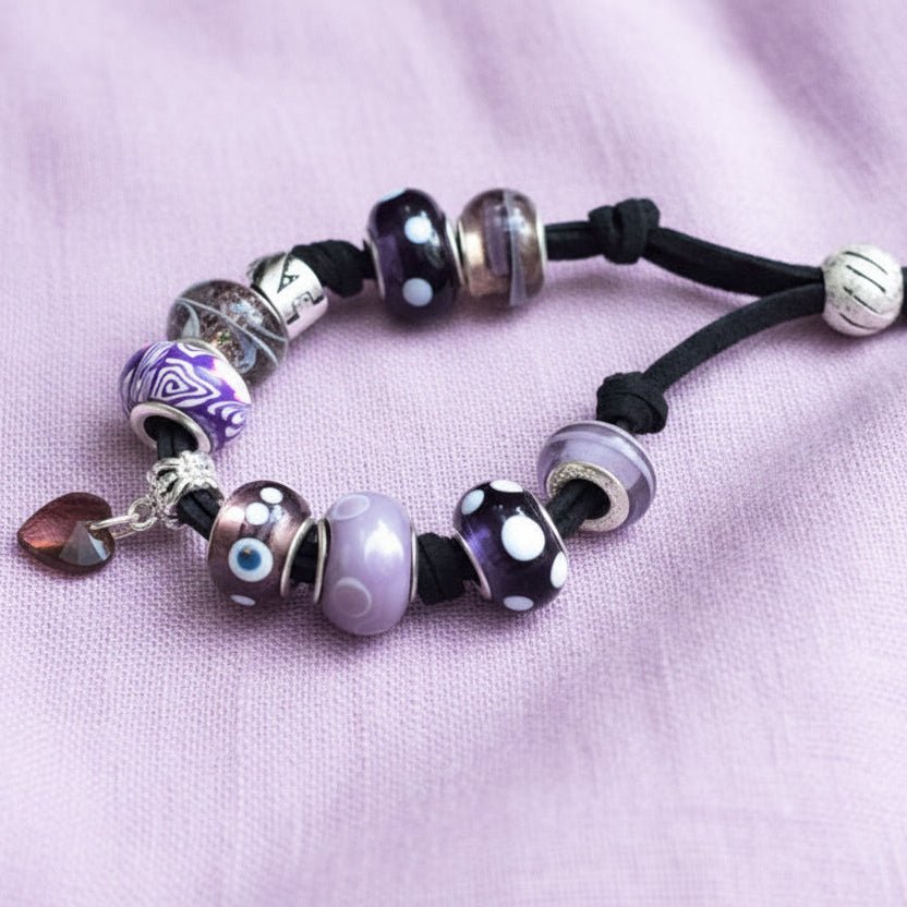 A purple glass charm bracelet with various beads and charms, including a purple heart-shaped Czech glass bead, and silver metal accents, strung on a black suede strap.