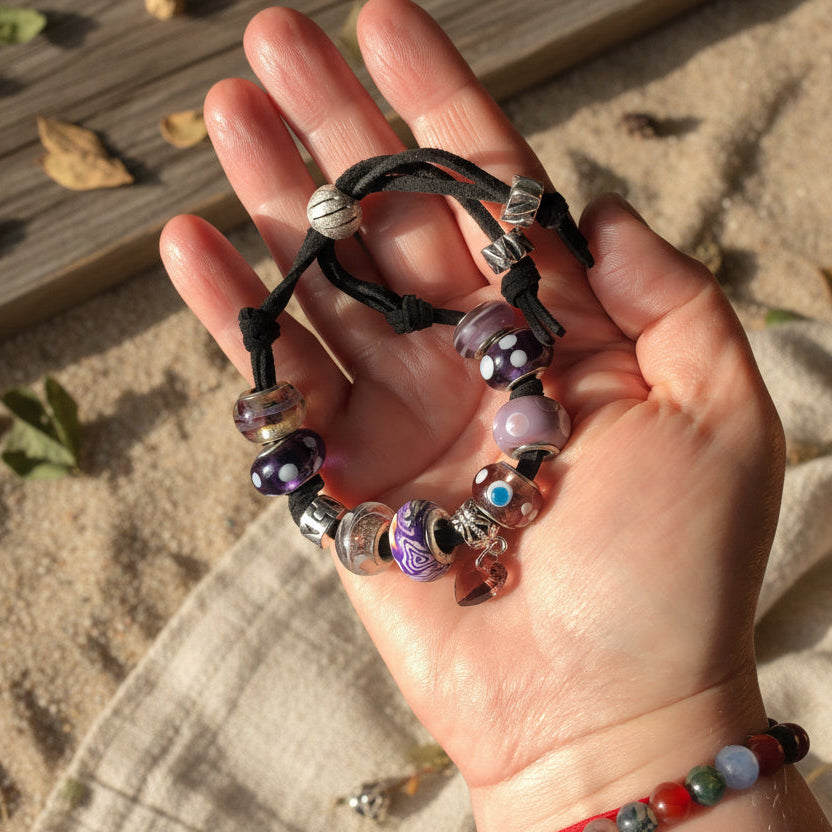 A purple glass charm bracelet with various beads and charms, including a purple heart-shaped Czech glass bead, and silver metal accents, strung on a black suede strap.