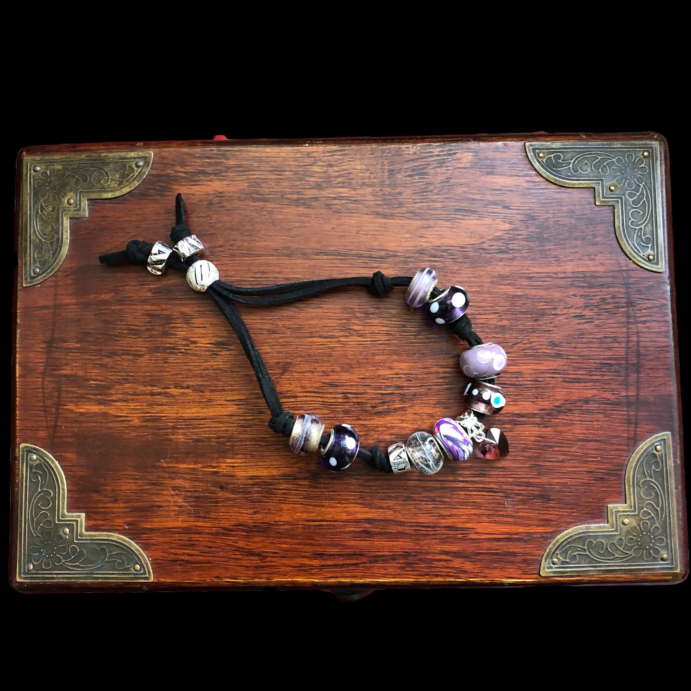 A purple glass charm bracelet with various beads and charms, including a purple heart-shaped Czech glass bead, and silver metal accents, strung on a black suede strap.